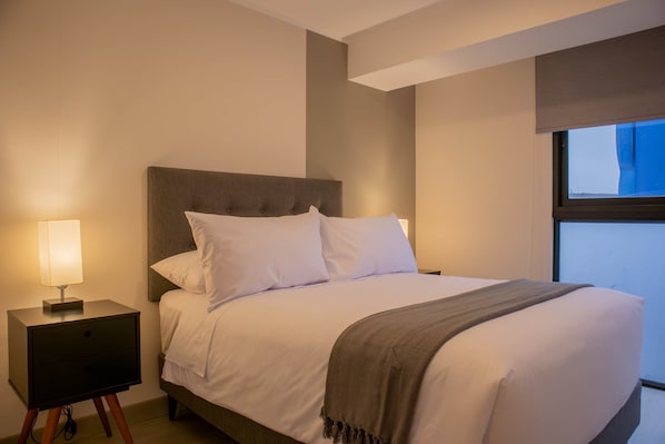 A neatly made bed with white linens and a brown throw blanket, a bedside lamp, and a nightstand with a drawer.