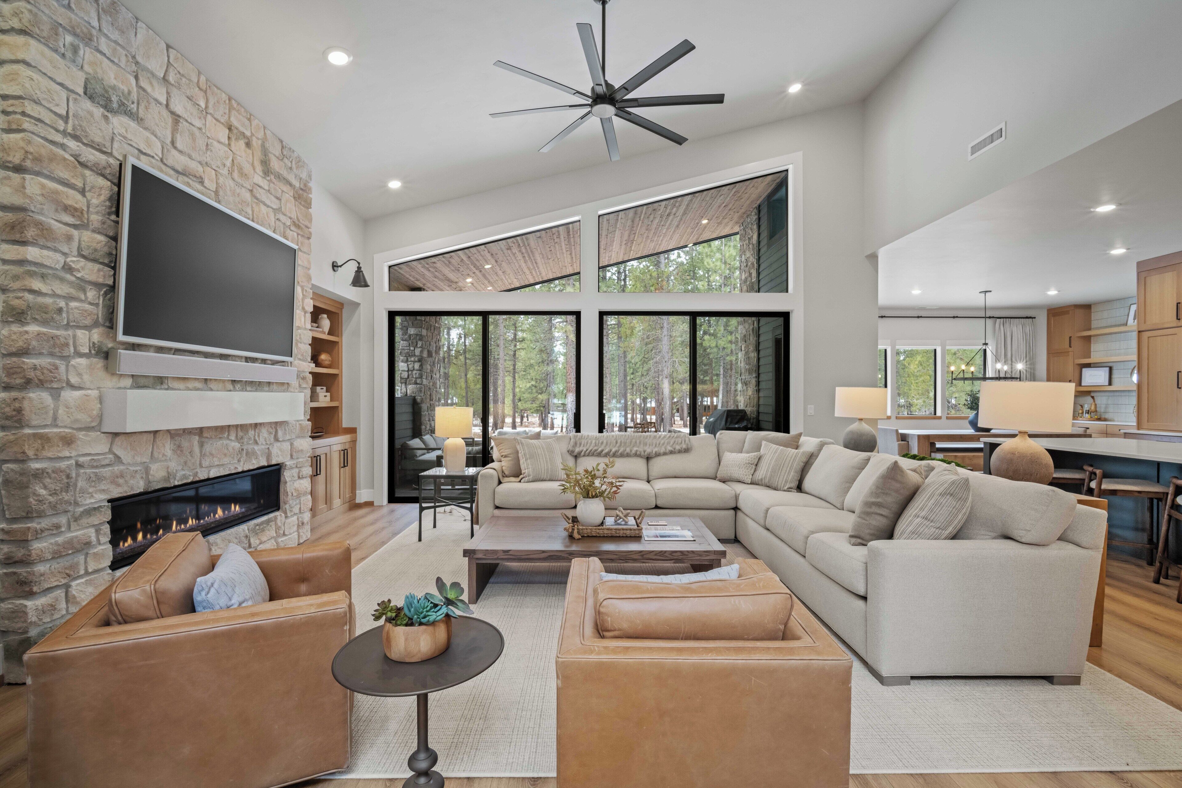 A spacious living room with a stone fireplace, large windows, a ceiling fan, and a modern kitchen in the background.