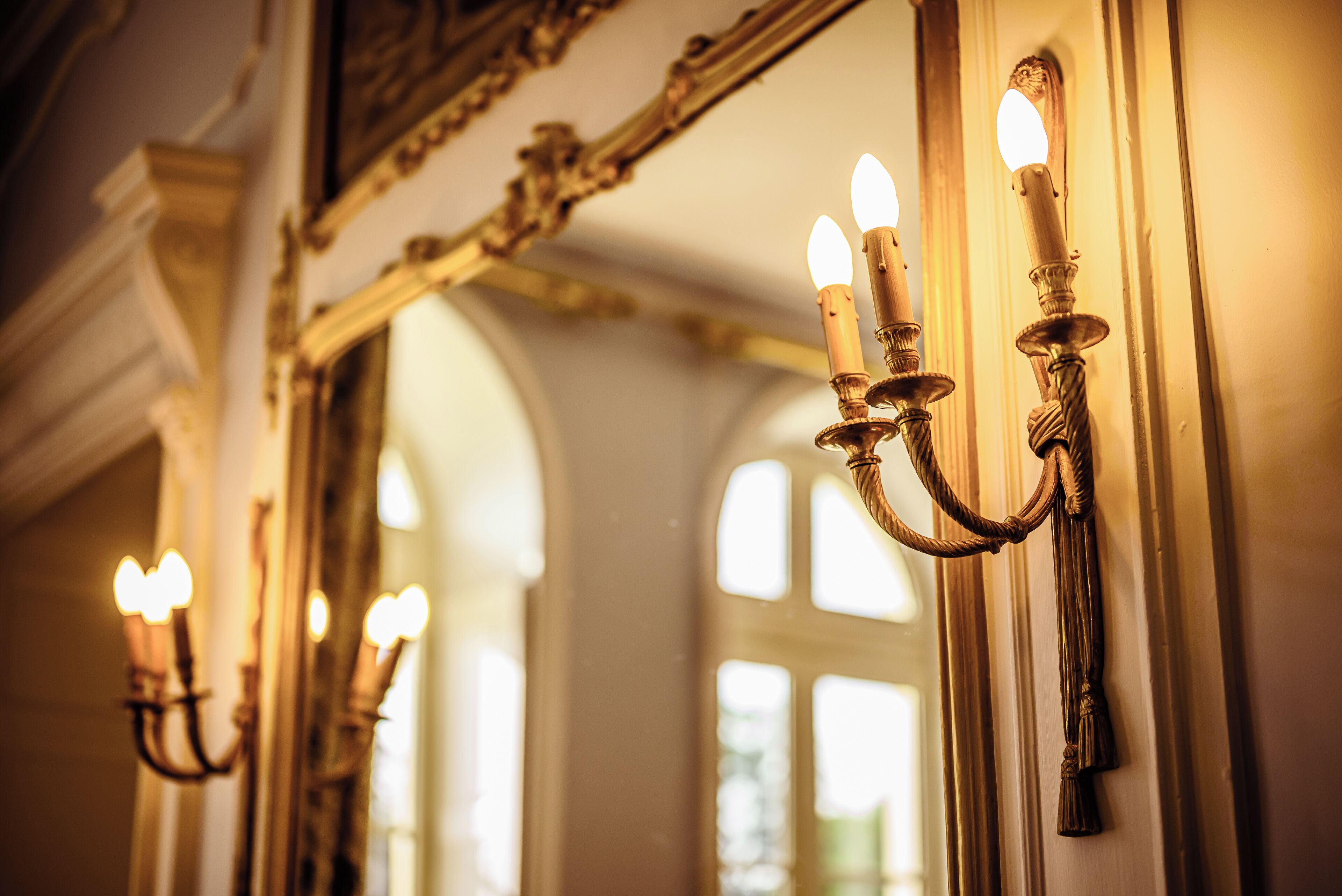 A corridor with ornate wall sconces and a mirror reflecting the interior.