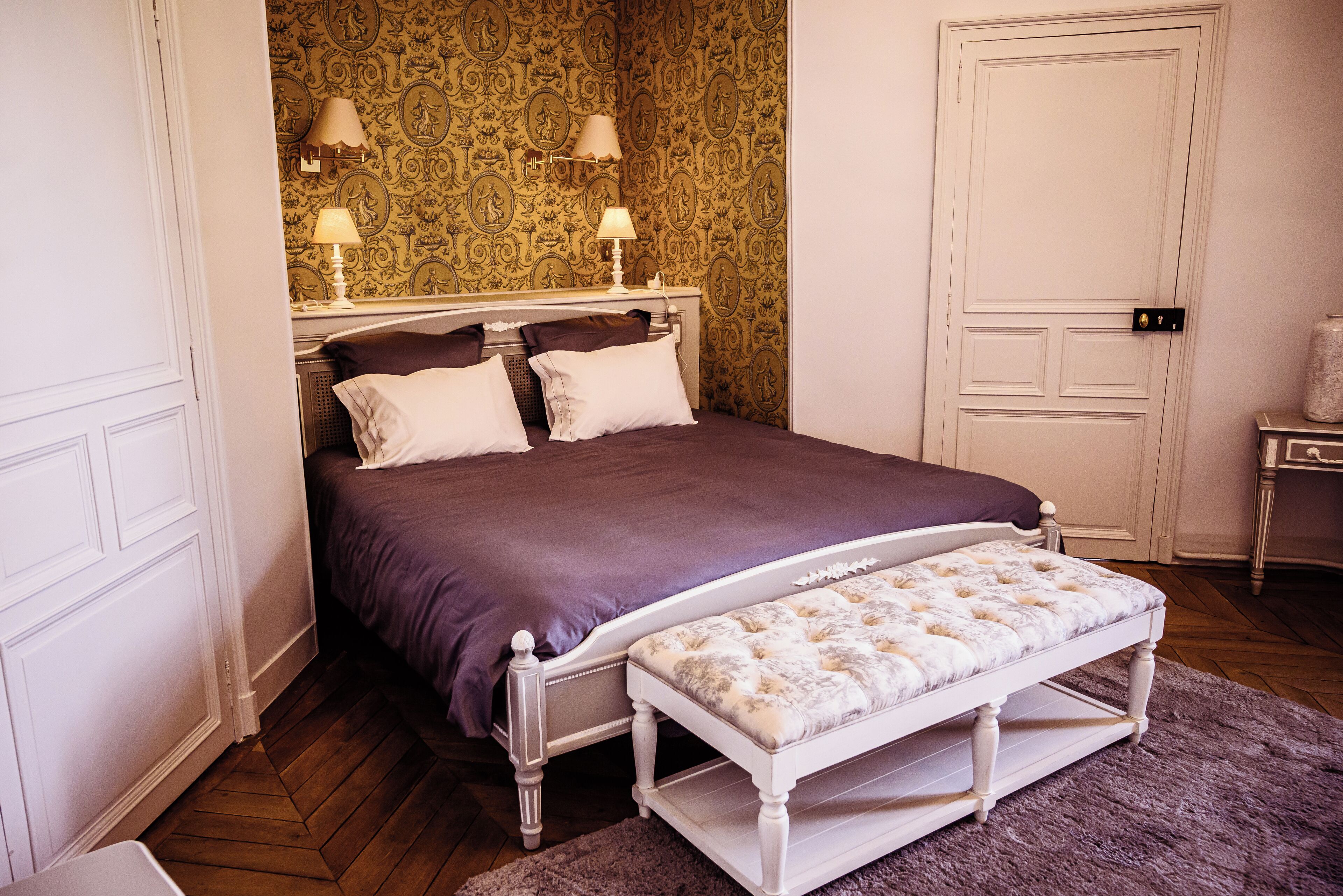A bedroom with a bed, two bedside lamps, a bench, and a door.