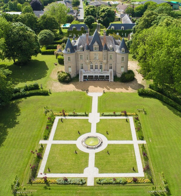 An aerial view of a large, historic building with a symmetrical garden layout, including a central fountain and manicured lawns.