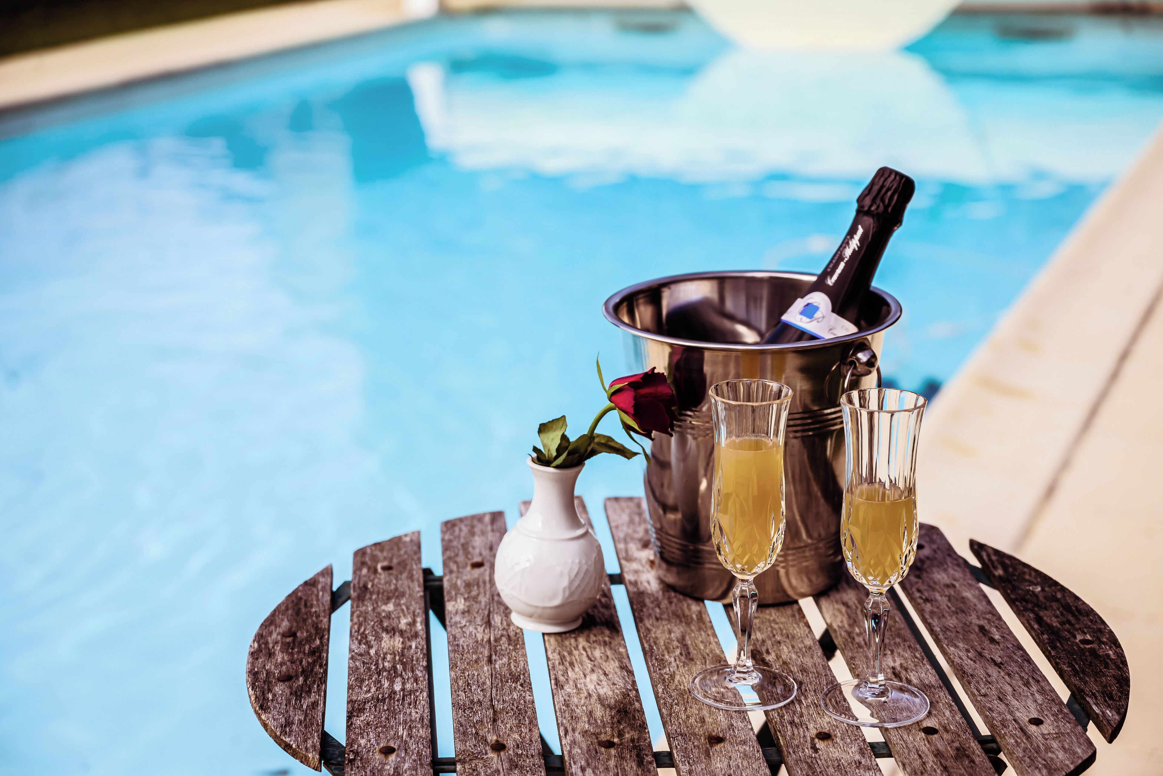 Una mesa de madera con un cubo metálico que contiene una botella y copas de champán, un jarrón blanco con una rosa roja, y una piscina al fondo.