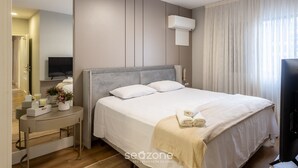 Una habitación de hotel moderna con una cama grande, una mesita de noche con lámpara, un televisor de pantalla plana y una ventana con cortinas.