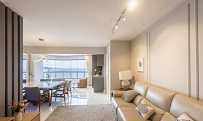 A modern living room with a sofa, a dining area with a table and chairs, and a view of the sea through large windows. - 3br Apt Sea View BBQ Garage - BC Vld1002 (Balneário Camboriú)