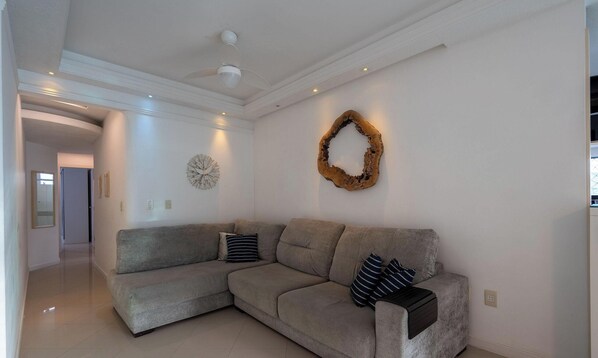 A modern living room with a grey sectional sofa, decorative wall art, and a ceiling fan. - Stay Steps From the sea in Meia Praia - VIM (Itapema)