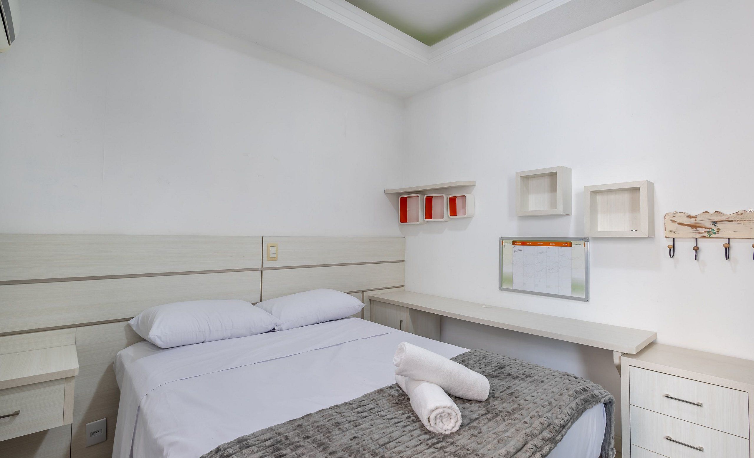 Quarto bem arrumado com uma cama, mesa de cabeceira, cômoda e prateleira na parede com caixas vermelhas e brancas.