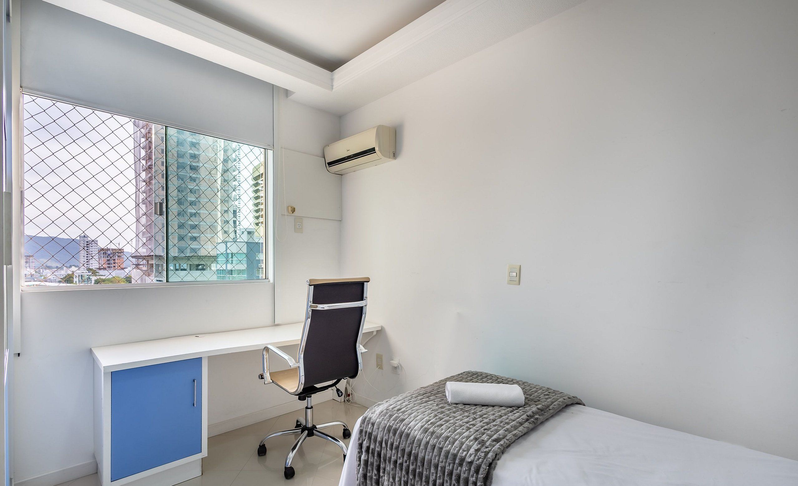 A modern bedroom with a bed, a desk with a chair, a window with a view of buildings, and an air conditioning unit.