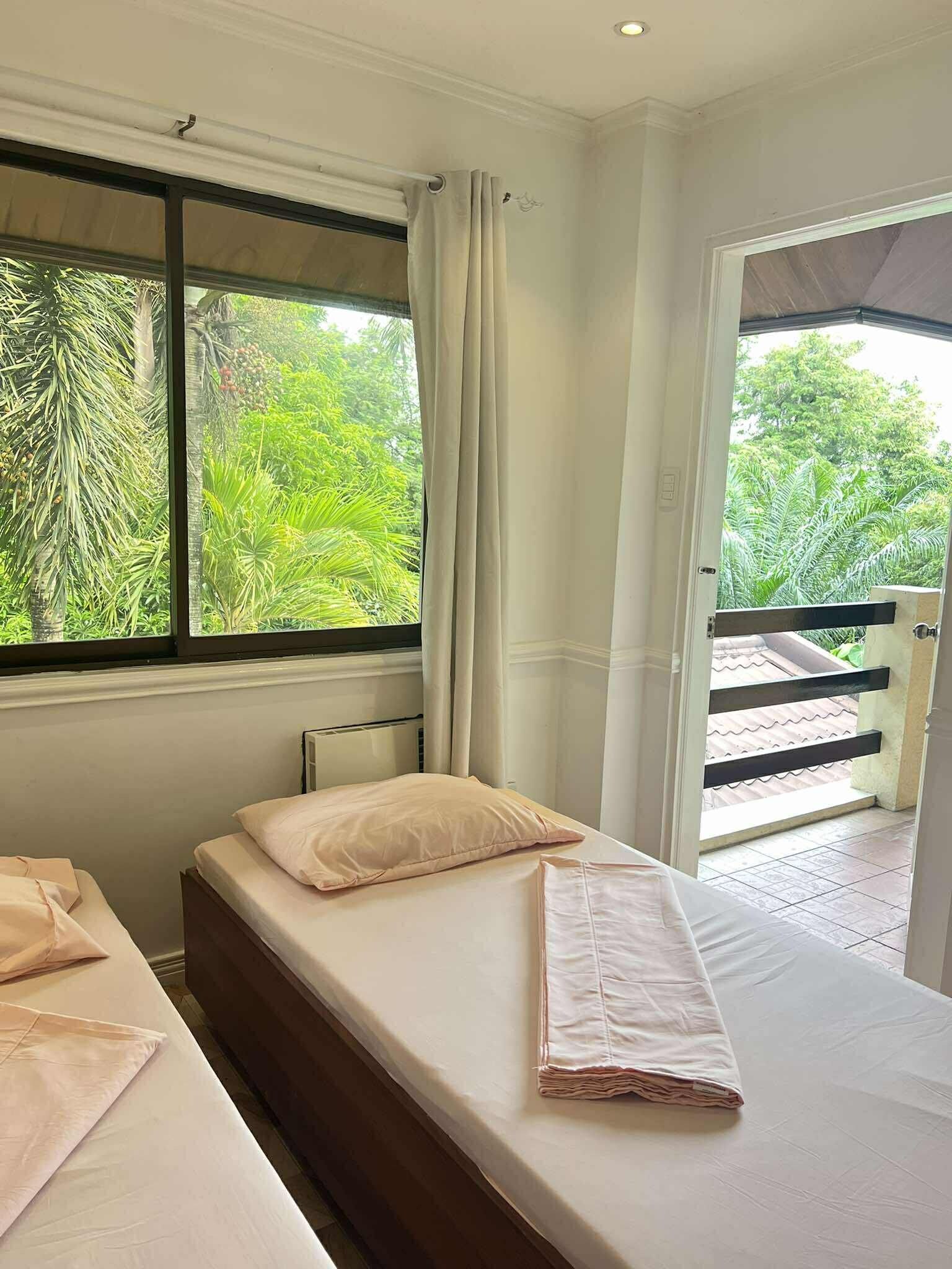 A room with two beds, a window with curtains, and a view of greenery outside.
