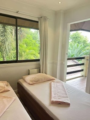 A room with two beds, a window with curtains, and a view of greenery outside.