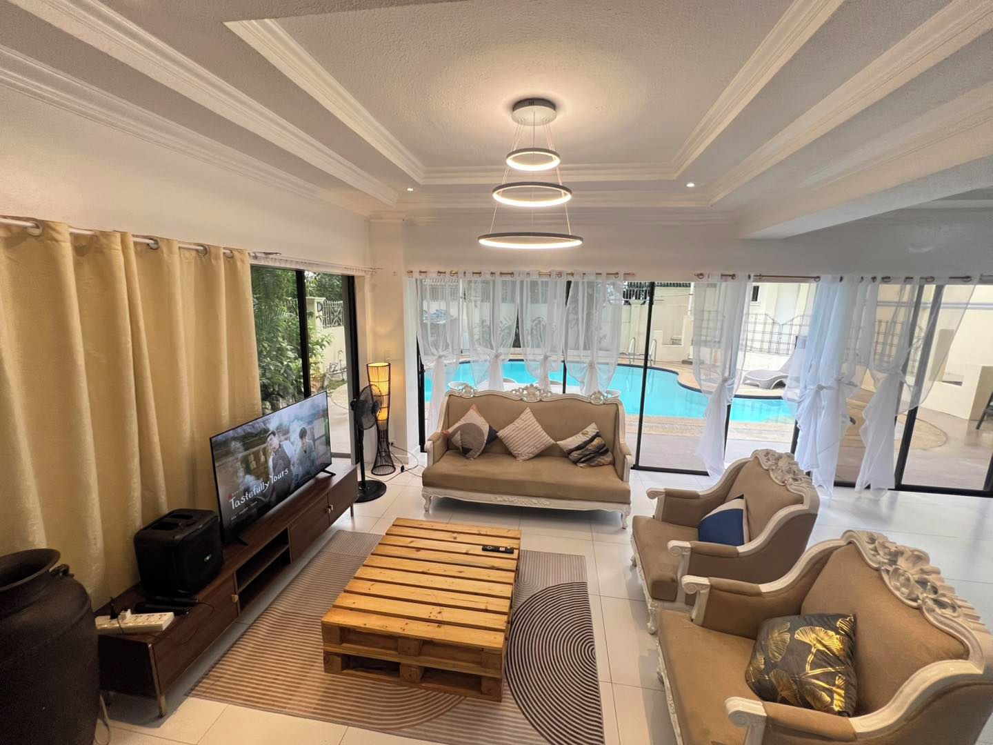 Amplia sala de estar con televisor, sofá y mesa de centro, vista a la piscina e iluminación empotrada en el techo.