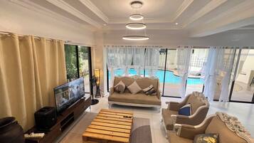 Amplia sala de estar con televisor, sofá y mesa de centro, vista a la piscina e iluminación empotrada en el techo.