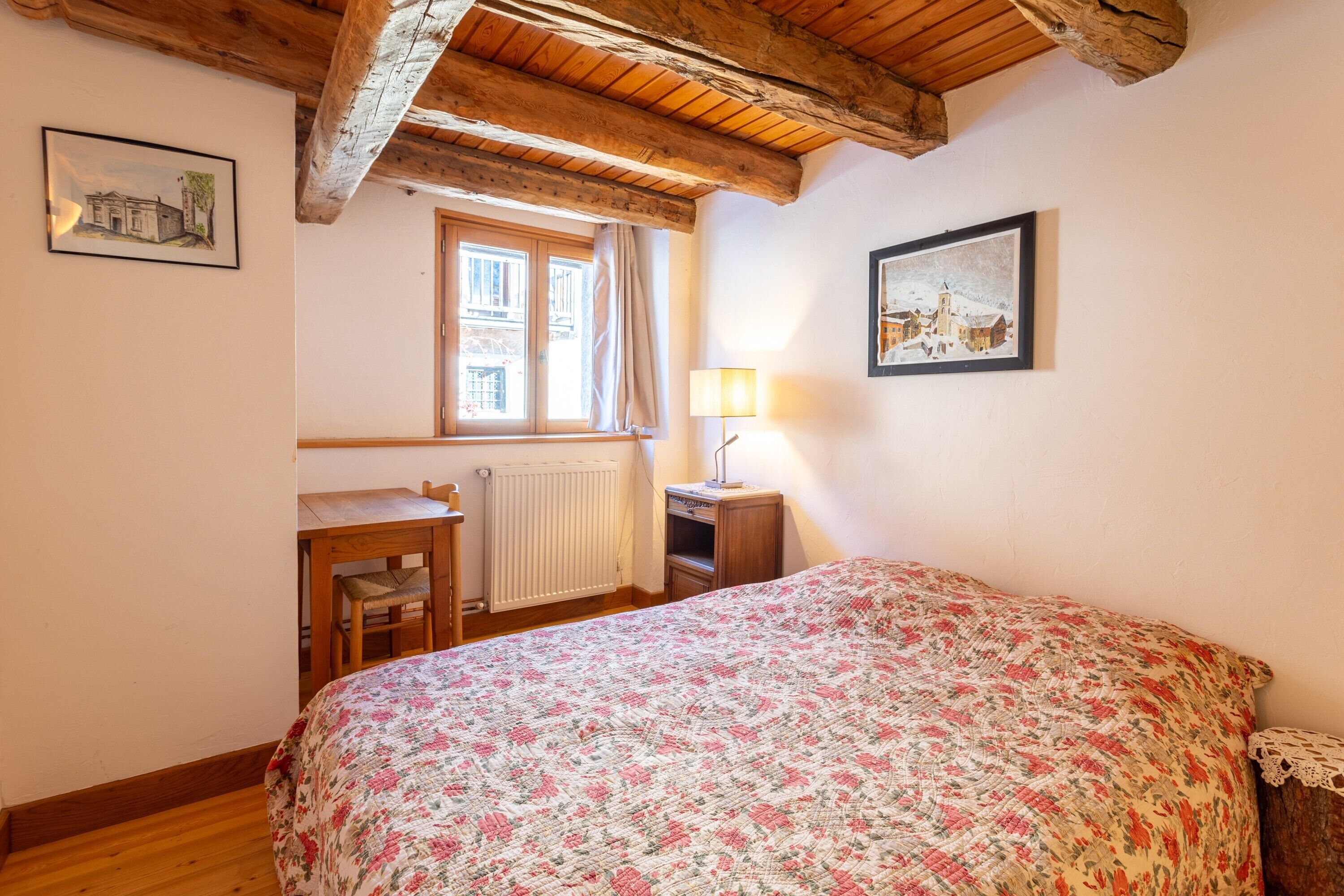A bedroom with a wooden ceiling, a bed with a floral bedspread, a desk with a chair, a window with curtains, and a lamp on a nightstand.