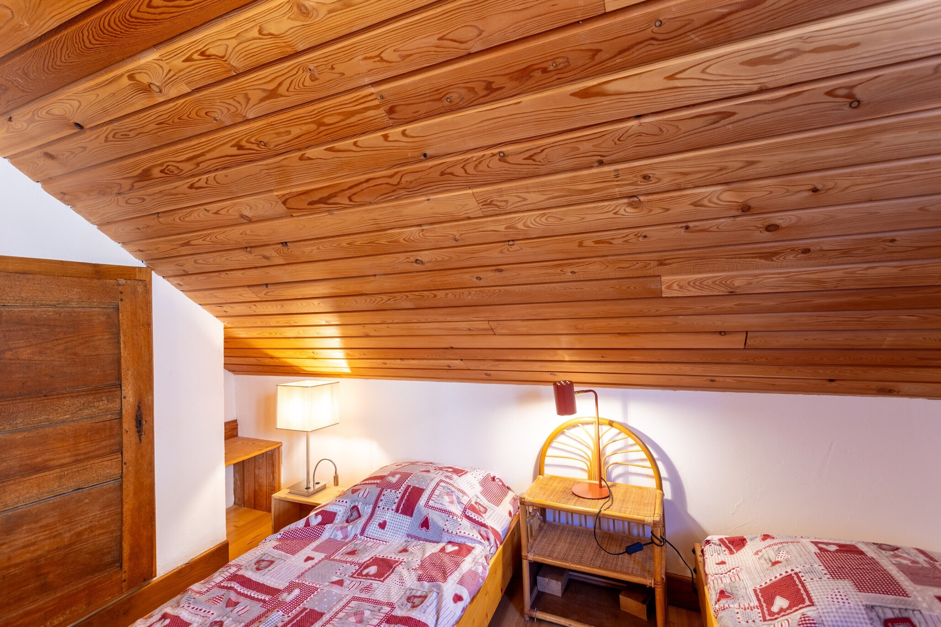 Techo de madera, dos camas con ropa de cama estampada, una lámpara y una silla.