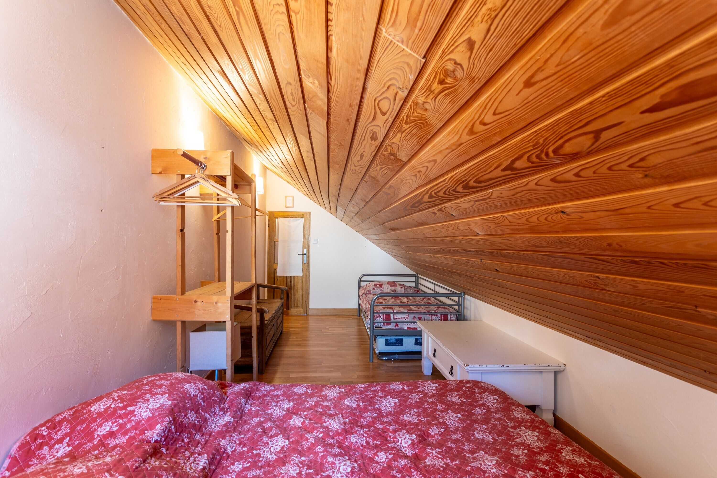 A small, cozy room with a sloped ceiling, a bed with a floral bedspread, a metal bunk bed, a wooden desk, and a hanging light fixture.