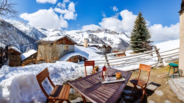 Een houten tafel met stoelen en een laptop, met een besneeuwd berglandschap op de achtergrond.