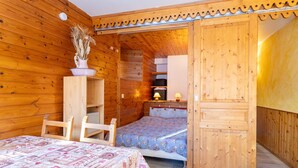 Een houten kamer met een bed, een stoel, een klein tafeltje met een lamp en een plank met decoratieve objecten.