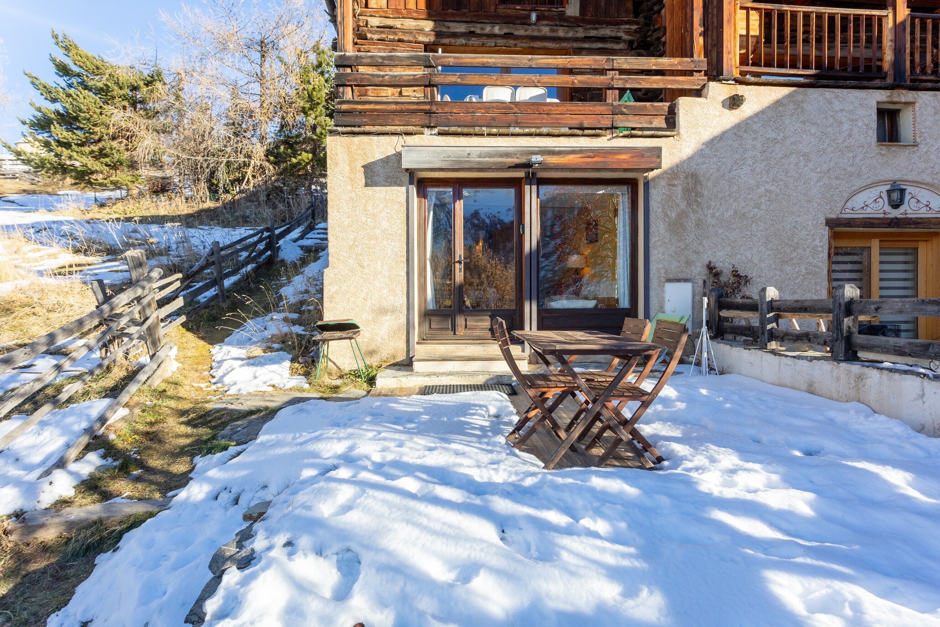 A chalet with a wooden balcony, a dining area with outdoor furniture, and a snow-covered ground.