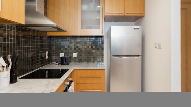 A modern kitchen with wooden cabinets, a stainless steel refrigerator, and a black tiled backsplash. - Serene 1BR at Iris Blue Marina (Dubai)