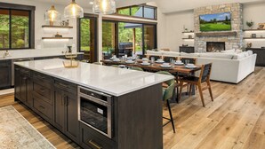 A modern kitchen with a large island, dark cabinetry, and stainless steel appliances. There is a dining area with a wooden table and chairs, and a living area with a stone fireplace and a large TV mounted on the wall. - Private Pool * Fantastic Outdoor Living in Suncadia! (Cle Elum)