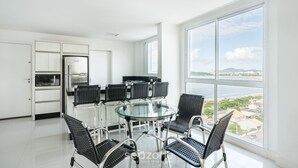 Una cocina moderna con armarios blancos y electrodomésticos de acero inoxidable, una mesa de comedor de vidrio con sillas negras y un amplio ventanal con vistas al mar y al paisaje urbano.