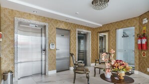 Un vestíbulo de hotel con tres ascensores, un mostrador de recepción, una silla, una mesa con un jarrón de flores y una araña.