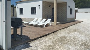 Un petit bâtiment blanc avec un toit en tuiles, une cheminée et un patio couvert comprenant des chaises longues blanches et un barbecue.