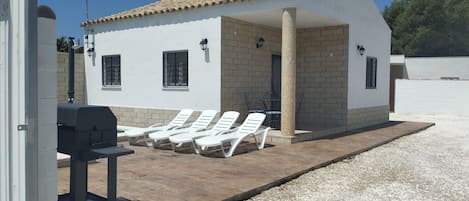 A small white building with a tiled roof, a chimney, and a covered patio area with white lounge chairs and a barbecue grill.