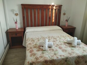 Una cama bien hecha con colcha floral, cabecera de madera, mesitas de noche con lámparas y un pequeño jarrón con flores en cada una.