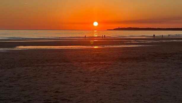Une plage au coucher du soleil, avec le soleil bas à l’horizon, des personnes se promenant le long du rivage et une côte lointaine parsemée de constructions.