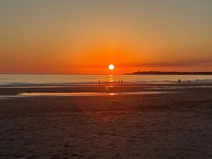 Una playa al atardecer con el sol bajo en el horizonte, personas caminando por la orilla y una costa distante con edificaciones.