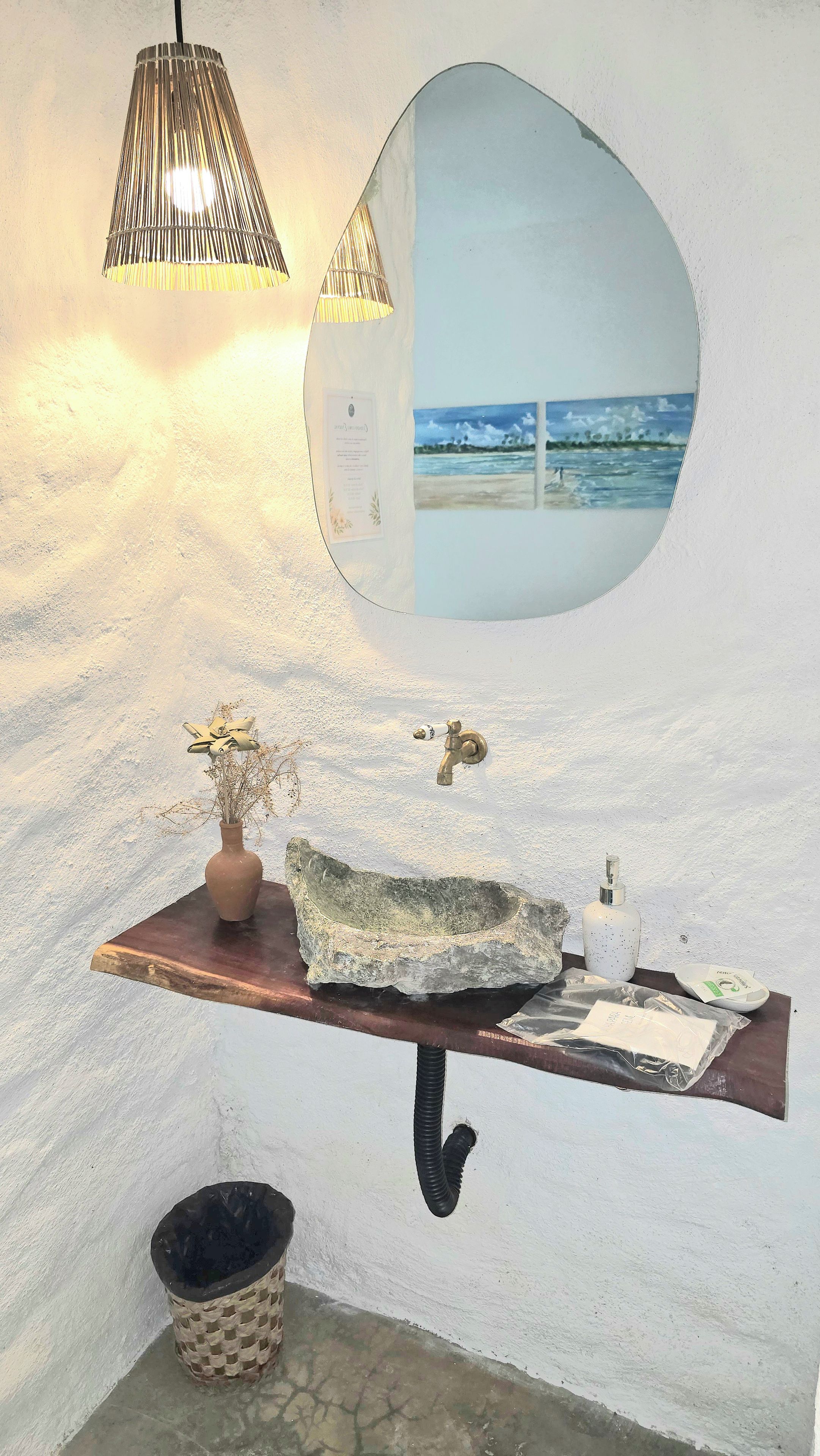 A bathroom with a round mirror, a wooden vanity, a hanging light fixture, a small trash bin, and a stone sink.