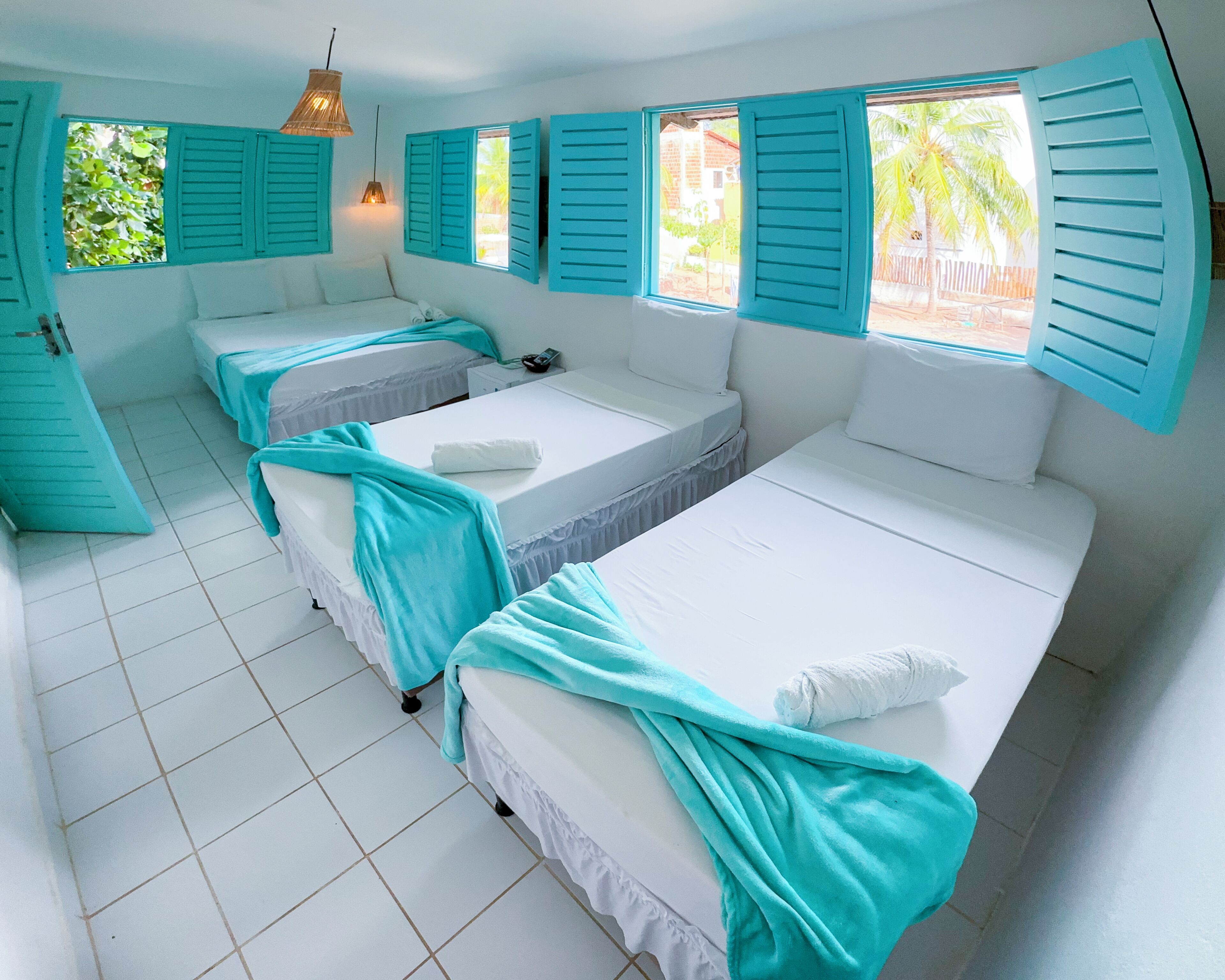 A room with two beds, each with a white mattress and teal bedding. There is a window with teal shutters, a hanging light fixture, and a tiled floor.