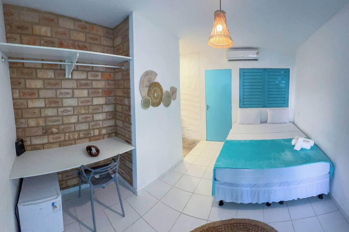 A room with a brick wall, a bed with a blue headboard, a white desk with a chair, and a hanging light fixture.