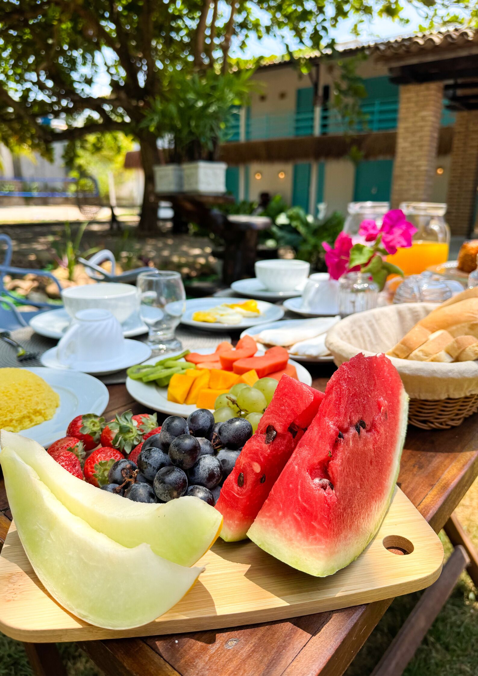 Una mesa de picnic con varias frutas y pan, rodeada por un edificio con tejado de tejas y vegetación.
