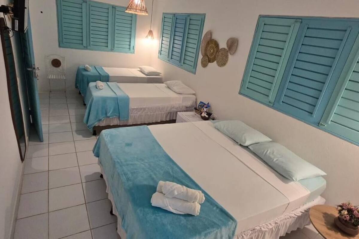 A room with three beds, each with a blue bedspread and white pillows, a small table with a vase of flowers, and a wall-mounted shelf with decorative items.
