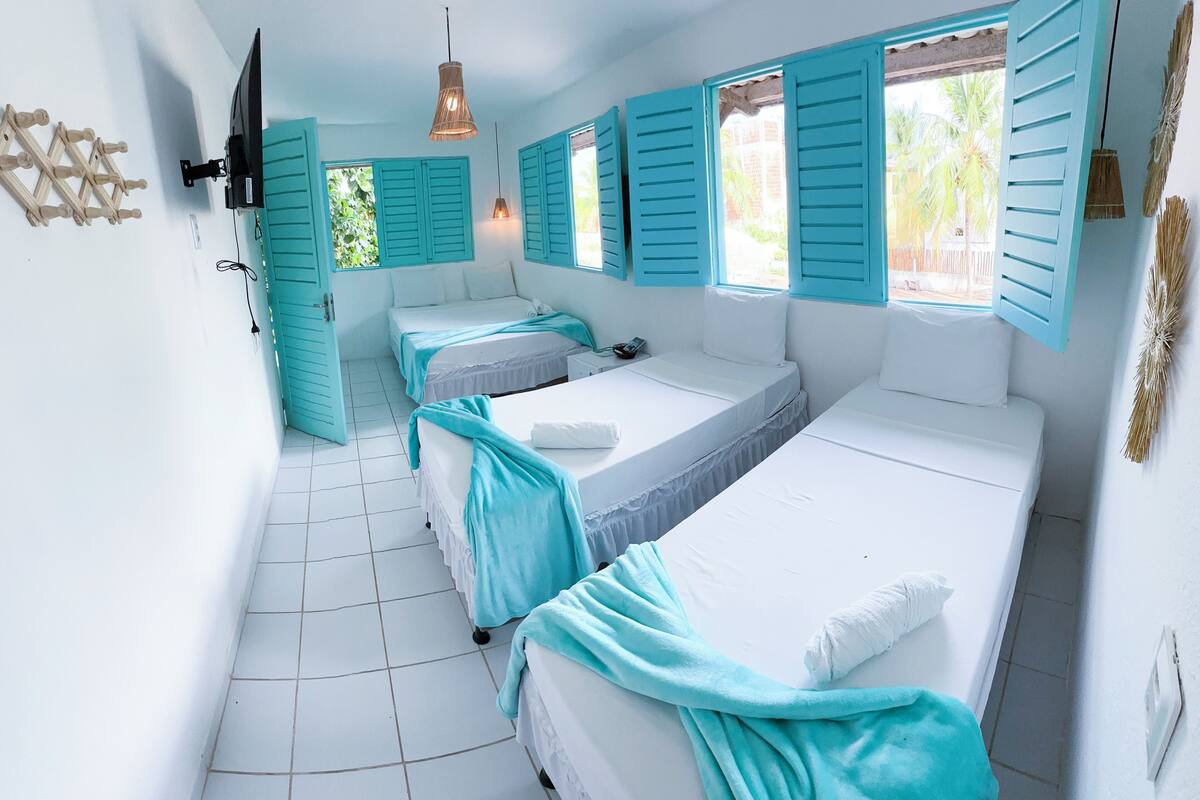 A room with three beds, each with white bedding and teal accents. There is a wall-mounted TV and a hanging light fixture.