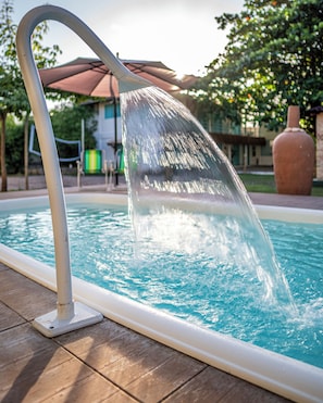 A pool with a water feature, a wooden deck, and a building in the background. - Amaly Pousada (Japaratinga)