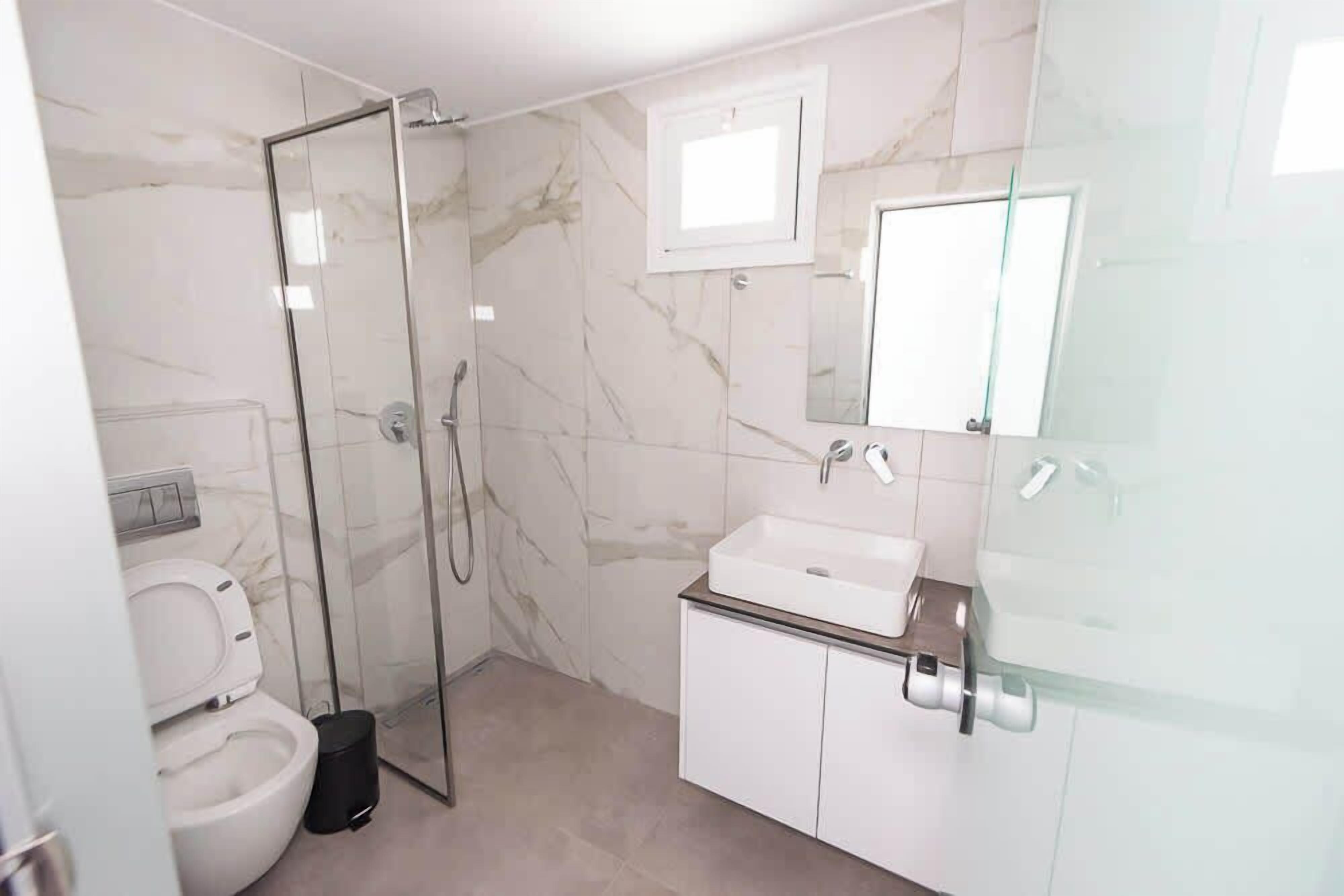 A bathroom with a glass shower enclosure, a white toilet, a sink with a mirror, and marble walls.