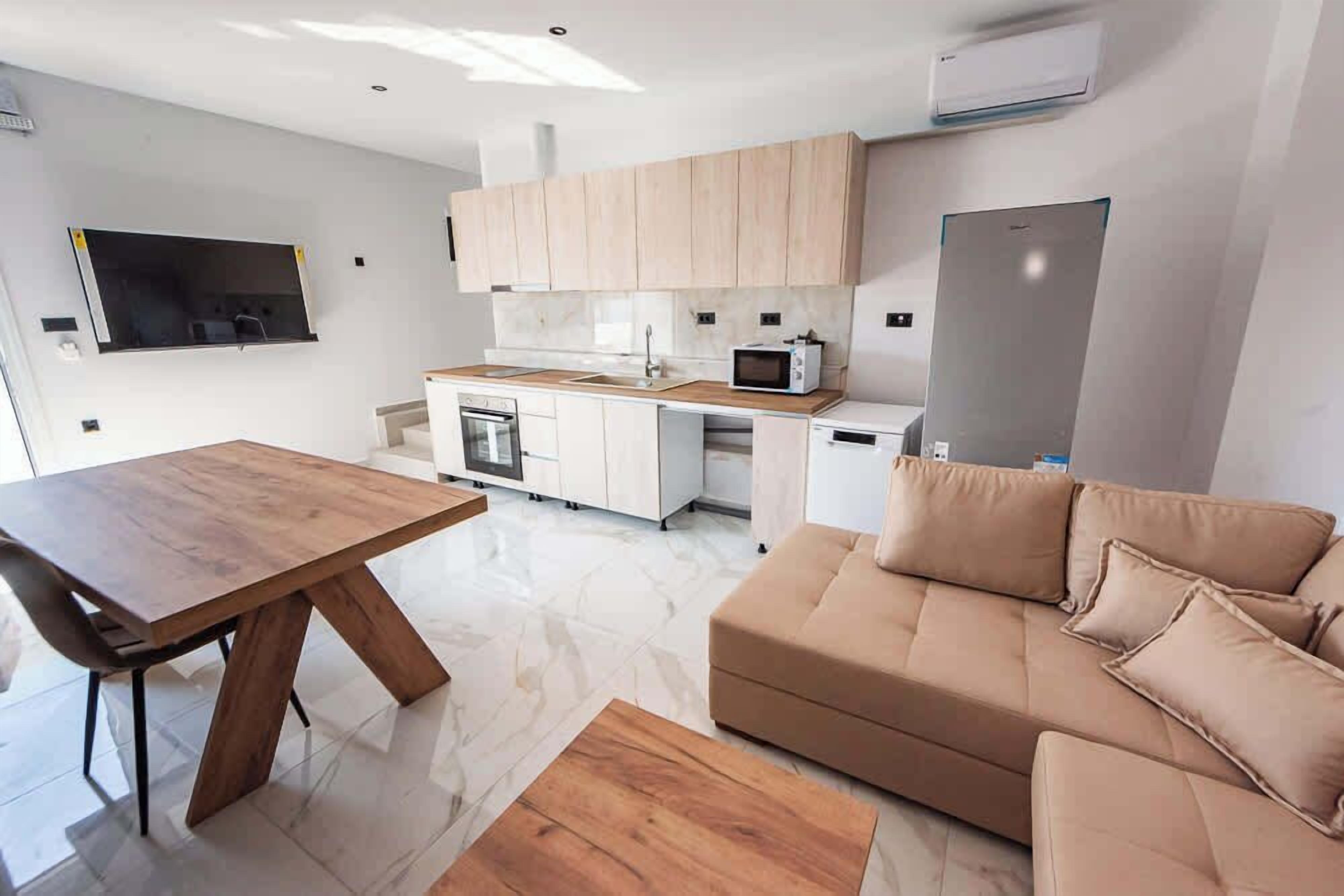A modern kitchen with white cabinets, a microwave, and a built-in oven. There is a wooden dining table and a beige sofa in the living area.