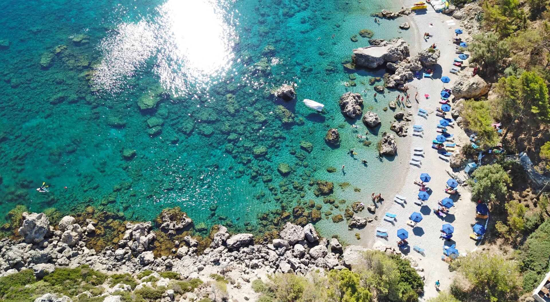 Vista aérea de una playa con agua turquesa cristalina, costa rocosa y sombrillas.