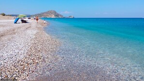 A pebbled beach with clear turquoise water, a distant rocky island, and a few people and umbrellas. - Sinani Villas Rhodes - Private Family Getaway (Afantou)
