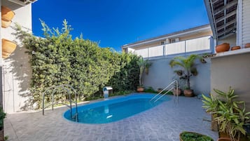 A swimming pool with a tiled deck, surrounded by potted plants and a hedge.