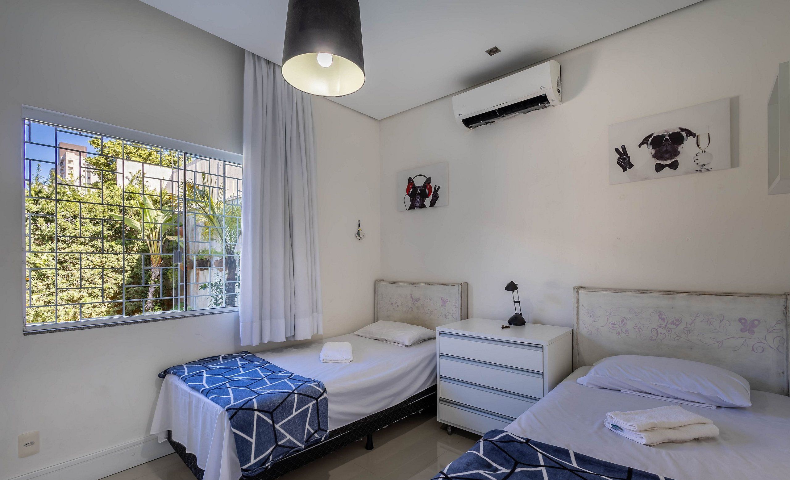 A bedroom with two beds, a window with a view of greenery, an air conditioning unit, and a wall-mounted artwork.
