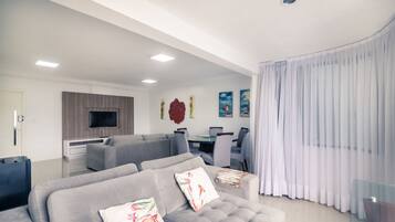 A modern living room with a grey sofa set, a flat-screen TV mounted on the wall, and a dining area with a glass table and chairs.