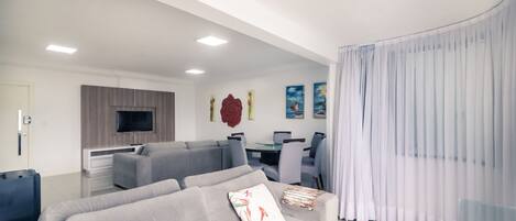 A modern living room with a grey sofa set, a flat-screen TV mounted on the wall, and a dining area with a glass table and chairs.