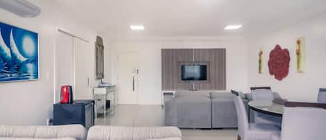 A modern living room with a grey sofa, a dining table with chairs, a flat-screen TV mounted on the wall, and a kitchen area in the background.