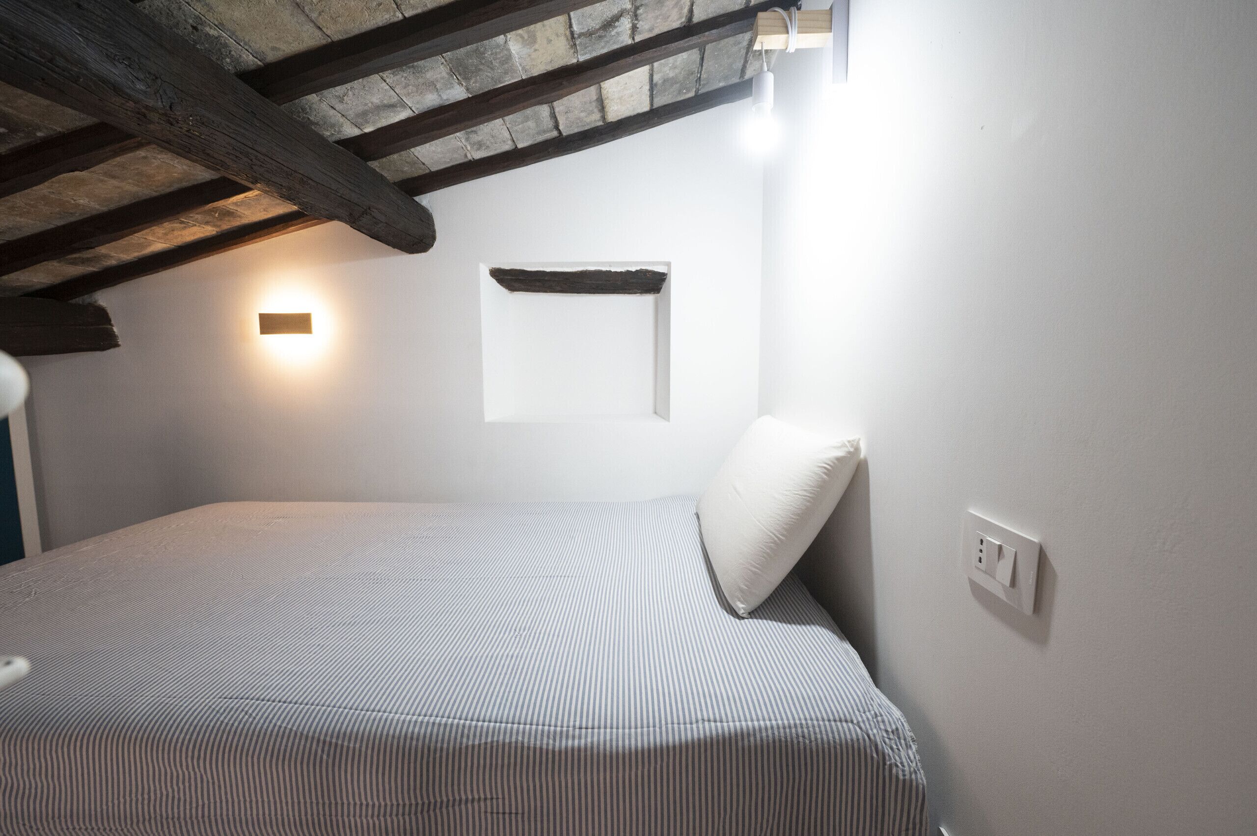 A bedroom with a bed, a wall-mounted light, and a wooden beam ceiling.