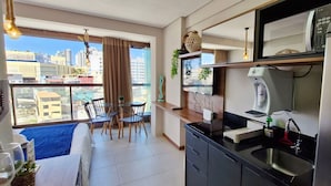 A modern kitchen with a stainless steel refrigerator, a built-in oven, and a sink. There is a dining area with a table and chairs, and a large window offering a city view. - COBAR0138 - Edifício Barra Premium (Salvador)
