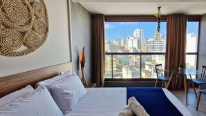A hotel room with a large bed, a dining table, and chairs. There is a view of a cityscape through the window. - COBAR0138 - Edifício Barra Premium (Salvador)