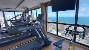 A gym with treadmills and a stationary bike, large windows with a city and ocean view. - COBAR0138 - Edifício Barra Premium (Salvador)